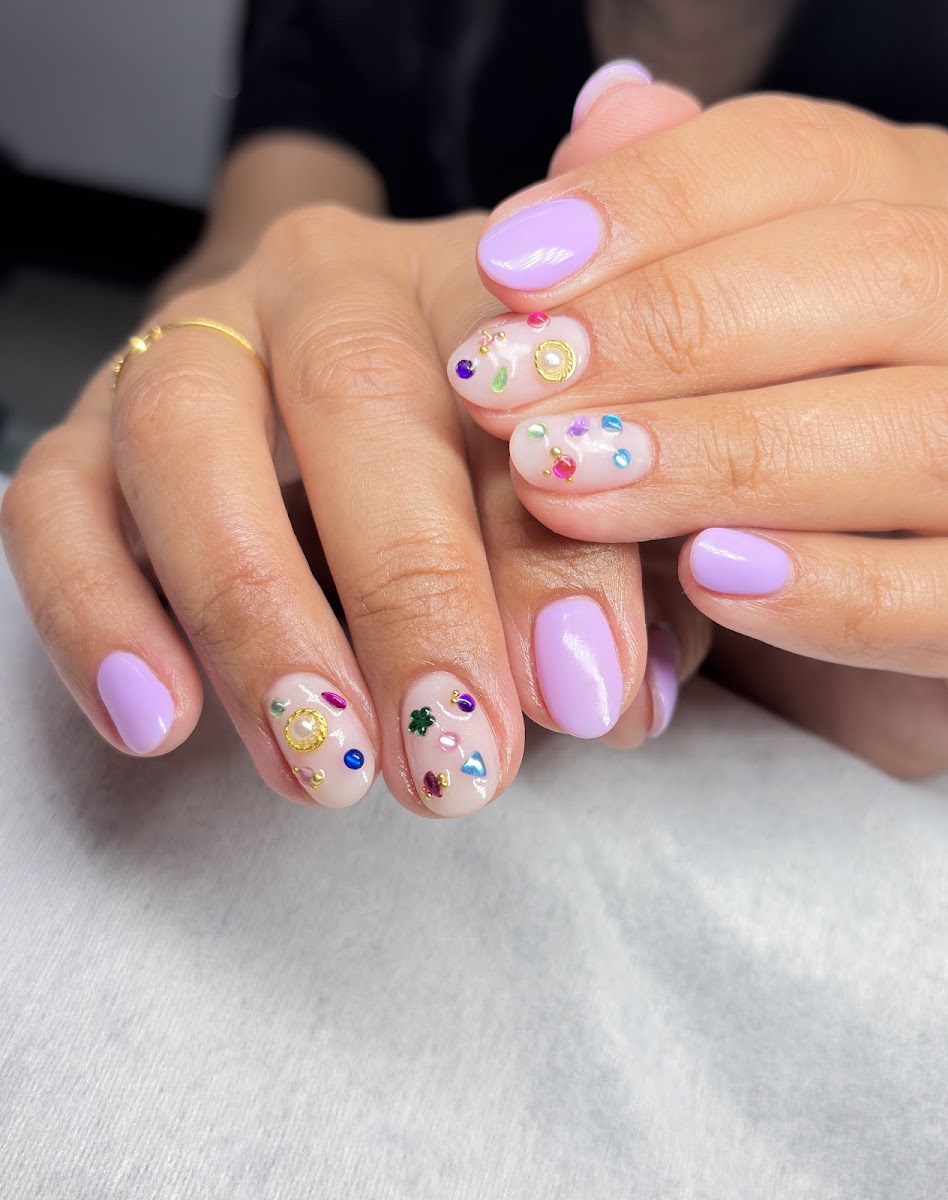 Manicura con uñas lilas y decoraciones de gemas de colores sobre base nude, manos con anillos dorados